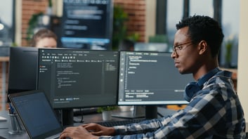 sofware-developer-thinking-while-touching-beard-while-typing-laptop-sitting-desk-with-multiple-screens-parsing-code-focused-database-admin-working-with-team-coding-background